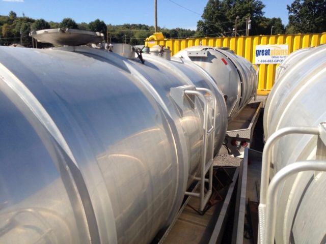 Vacuum tanker trailer cylindrical metal tank with access hatches and ladders, parked outdoors with other tanks and yellow barriers in the background.