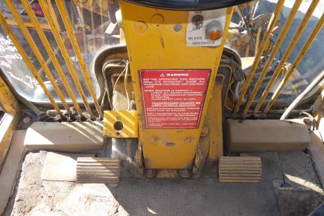 Caterpillar 16G grader foot pedals and central control panel with warning label and hydraulic lines visible.
