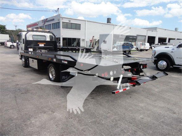 Isuzu NRR tow truck with flatbed body in an outdoor lot, stock number NRRDSL22XLP visible on building, other trucks nearby.