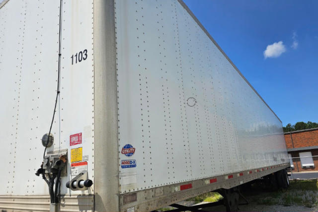 Utility trailer side view showing 1103 number, riveted aluminum panels, and rear doors. Sticker labels near front corner and a blue Utility 4000-D-X badge visible.