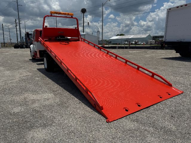 Tow truck with a bright red flatbed ramp extended, I-beam side rails, and an orange beacon light on top of cab.