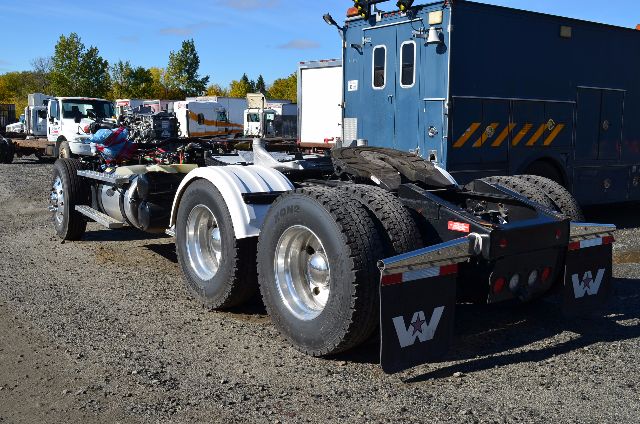Truck chassis with dual rear axles and mud flaps showing Western Star logos. Blue cab-over in background. Visible text on mud flaps: W and decorative stripes.