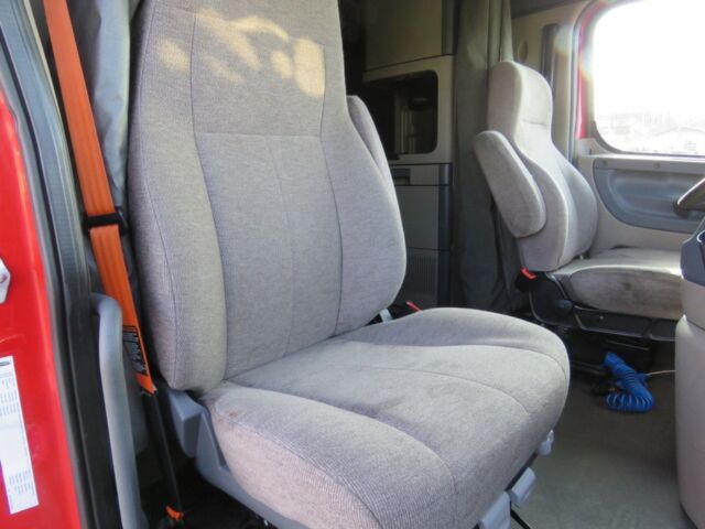 Cabin interior showing front seats of a Freightliner Cascadia Evolution truck, light gray fabric upholstery, steering wheel visible, driver seat with seatbelt