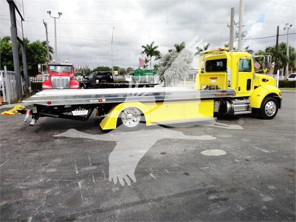 Tow truck with a bright yellow Peterbilt 337 and a long flatbed trailer in a dealership lot.