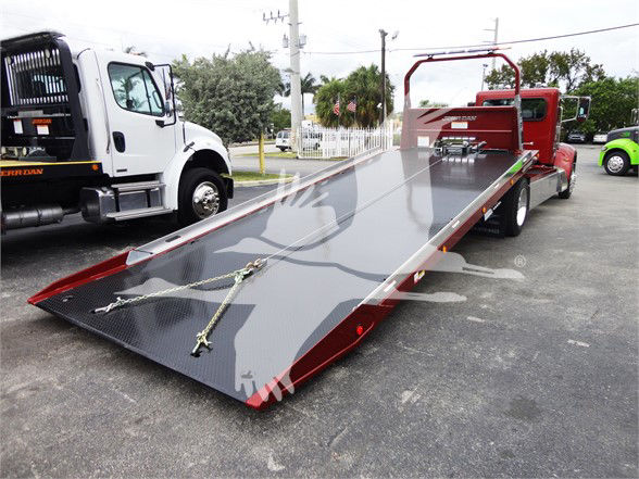 Peterbilt 337 flatbed tow truck in lot, large metallic ramp with tiedowns, red cab, white tow truck on left.