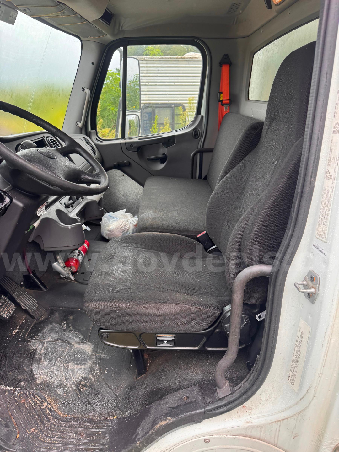 Cab interior of a 2016 Freightliner M2 106 truck showing driver and passenger seats, steering wheel, dashboard, floor with dirt, and side door with window.