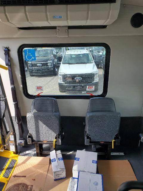 Ford E-450 shuttle bus interior with two gray seats, view through side window showing a white Ford SUV outside; boxes on the floor near seats.