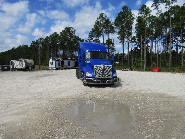Blue 2020 Kenworth T680 truck in a gravel lot with trees in the background, front grille and bumper visible.