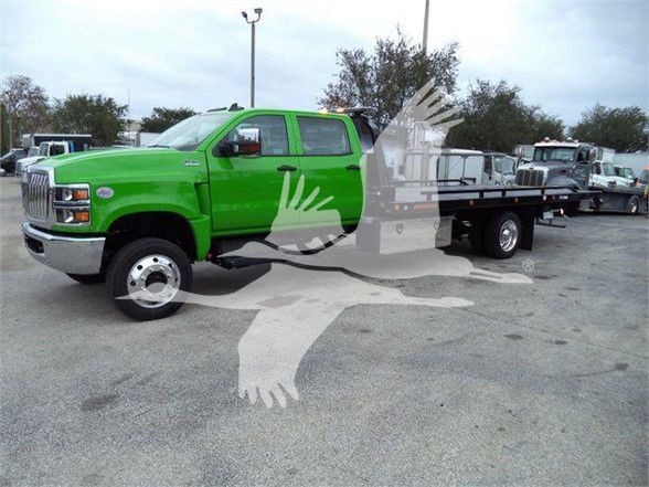 Green 2023 International CV tow truck with flatbed, cab shows GMC-like grille, side mirrors, and black flatbed behind. Visible text on vehicle not legible in image.