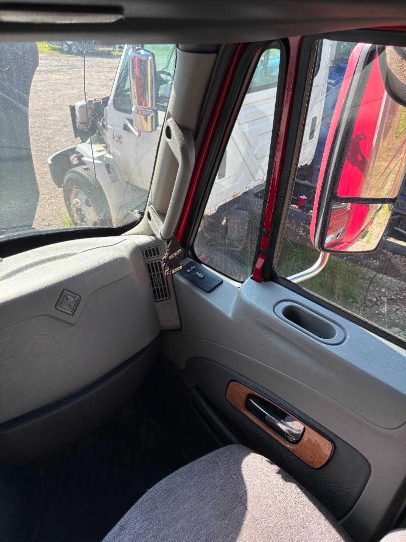Truck cab interior showing front passenger side with grey dashboard, vertical window pillar, wood trim on door, and side mirror; window reflections of another truck outside.