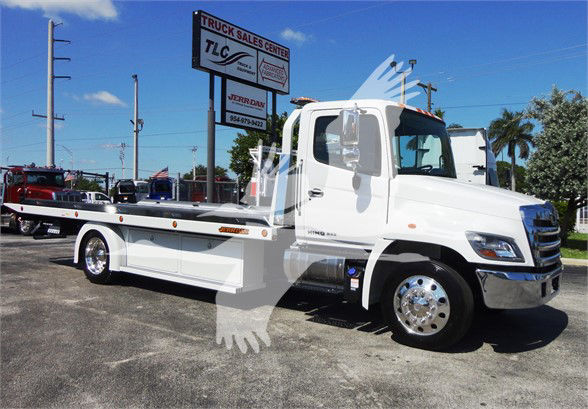White 2022 Hino 268A tow truck side view with flatbed, chrome wheels, and CDL cab. Signage reads TRUCK SALES CENTER in background.