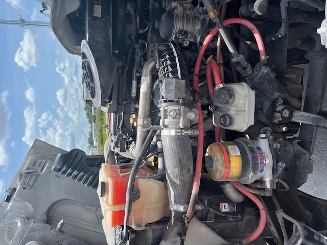 Engine bay of a Freightliner Cascadia with visible coolant reservoir, power steering fluid reservoir, and various red hoses and hoses in a diesel engine compartment.