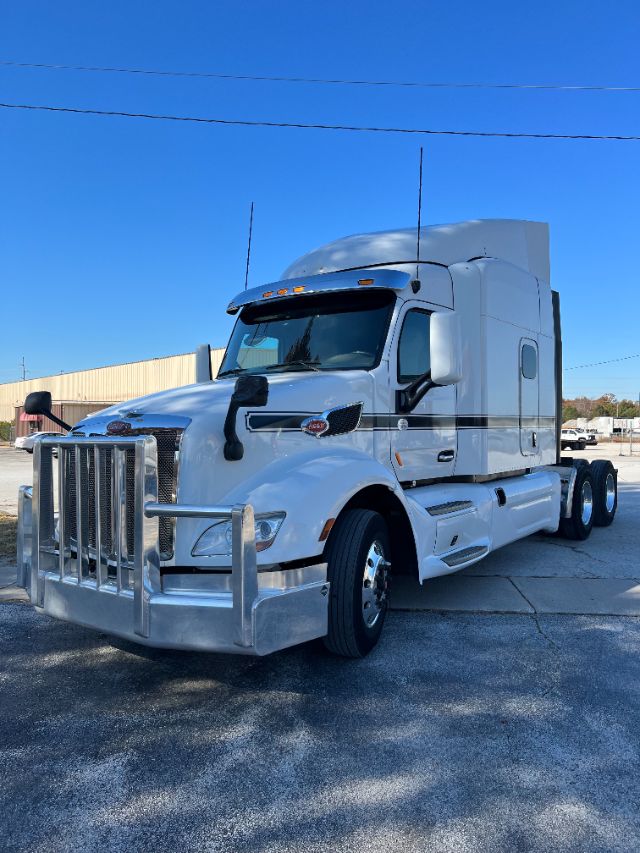 Peterbilt 579 truck in white, front grille guard, sleeper cab, tall exhausts, side mirrors, and dual rear axles in a parking lot.