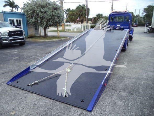 Tow truck deck on a 2025 Kenworth T280 with a long tiltbed trailer, blue truck in the background, hydraulic winch chains visible.