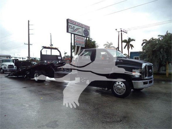 Ford F-750 SD tow truck with a car carrier trailer, visible text includes stock sign in background and dealership banners; no identifiable model text clearly readable on truck beyond general appearance.