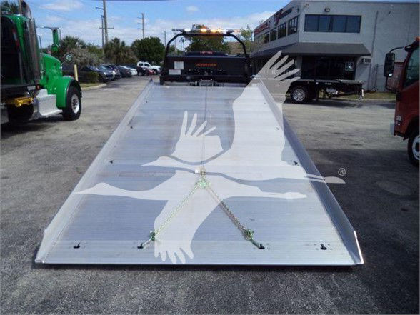 Tow truck platform with ramp and a bird graphic on aluminum surface; background shows other trucks and a building.