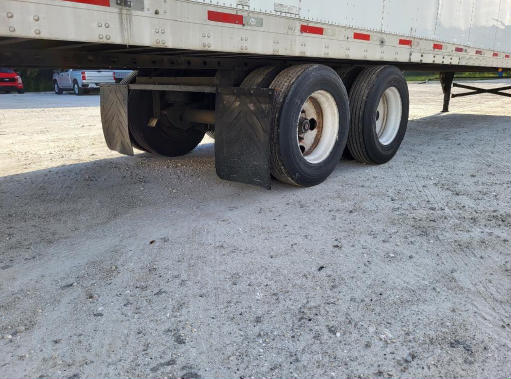 Trailer undercarriage showing dual rear axles with mud flaps and tires, on a gravel surface.