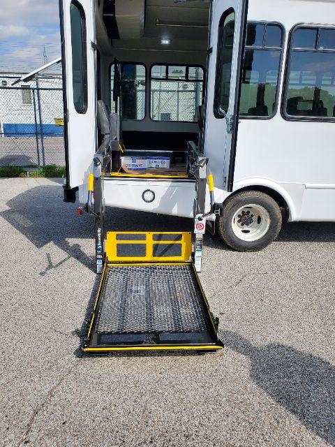 Ford E-450 shuttle bus with side doors open, wheelchair ramp deployed. Visible labels: 'TRANSIT BUS' on a box inside. Ramp platform with black grip surface and yellow edges. Front wheel and bus body visible.