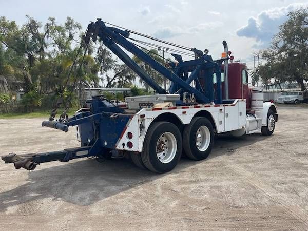 Rear and side view of a tow truck with a blue boom, white body, and red cab. No visible text legible on the truck.