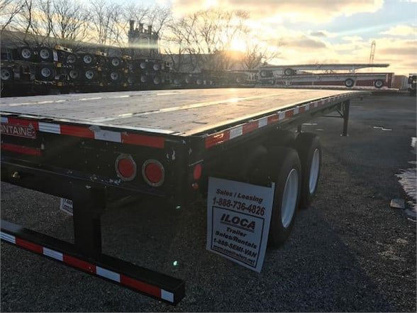 Flatbed trailer with dual axles and reflective striping, rear lights, and a hanging sign showing leasing information. Sunset over stacked trailers in background.
