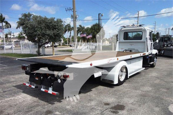 Flatbed tow truck rear/side view with wooden bed, white cab, and hydraulic controls visible.