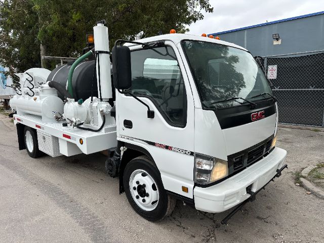 GMC W5500 vacuum truck with white cab and vacuum tank. Side shows 'GMC', '3500', and various hoses connected to the tank. Front grille clear, headlights, and orange warning lights on roof.