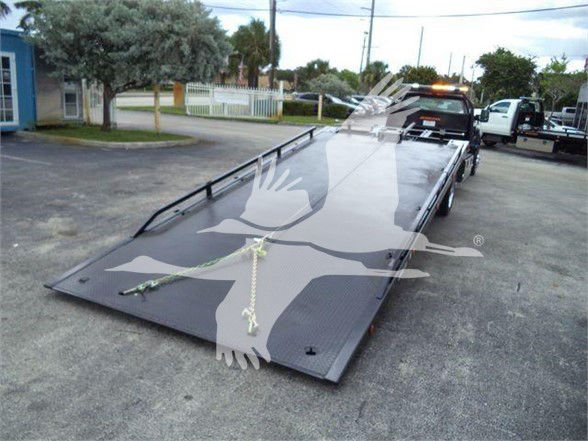 Tow truck flatbed trailer with hydraulic tilt showing a long, gray steel ramp on an asphalt lot.