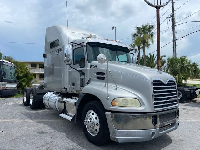 Truck cab of a 2010 Mack CXU613 conventional sleeper in a parking lot, front view with chrome bumper, grille, and tandem rear axles.