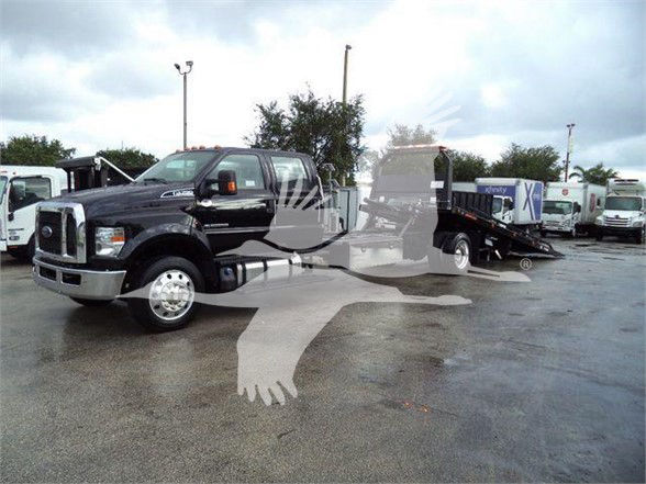 Tow truck 2024 Ford F-750 SD in a dealership lot, black crew cab with tow bed attached, other trucks in background.