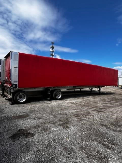 Flatbed trailer with rolling tarp parked on gravel, red tarp cover along trailer length.