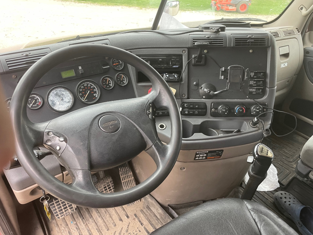 Truck interior showing steering wheel, gauge cluster, and center dashboard of a Freightliner Cascadia with a manual shifter.