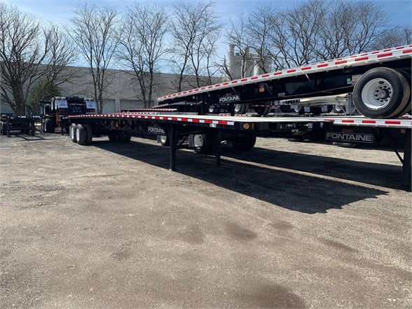 Flatbed trailer with a Fontaine Velocity steel trailer in a yard, long low-slung platform with reflective edge strips and dual rear axles.