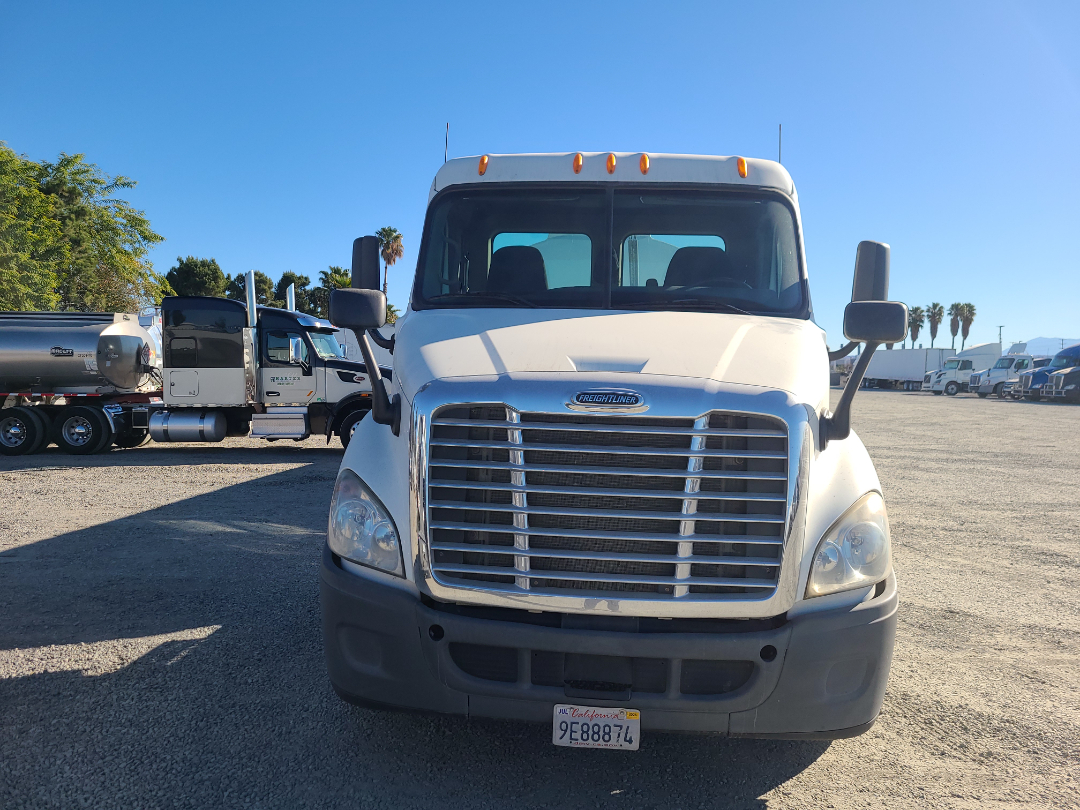 White Freightliner semi truck front view with large chrome grille, hood, and California license plate 9E887L4. Bright day, other trucks in background.