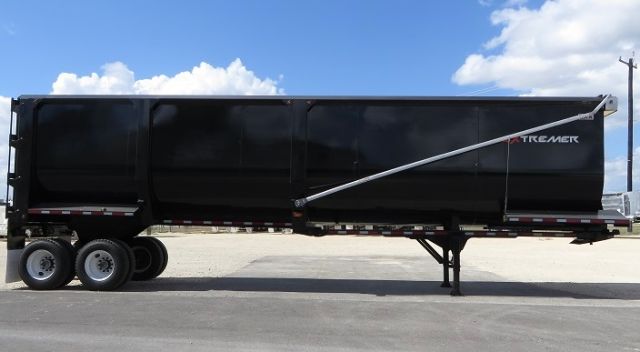 Tandem-axle dump trailer with frameless design and rear-mounted hydraulic hoist; black exterior with Tremor logo on front.