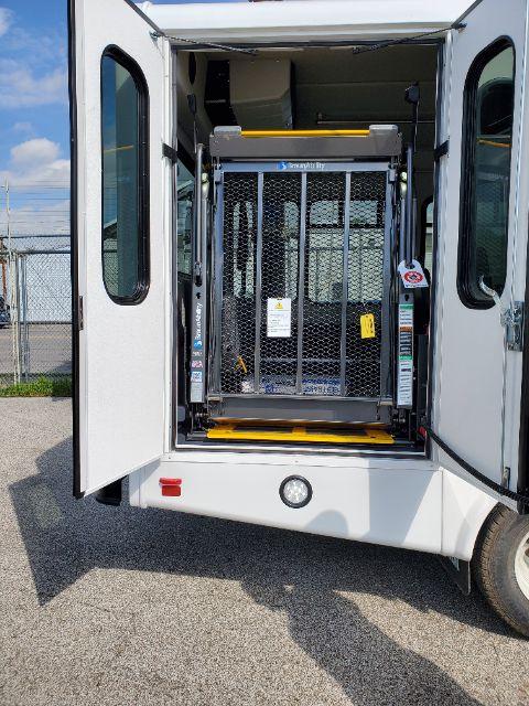 Front view of a 2025 Ford E450 shuttle bus with a wheelchair lift installed, rear doors open, showing the elevator platform inside the entryway.
