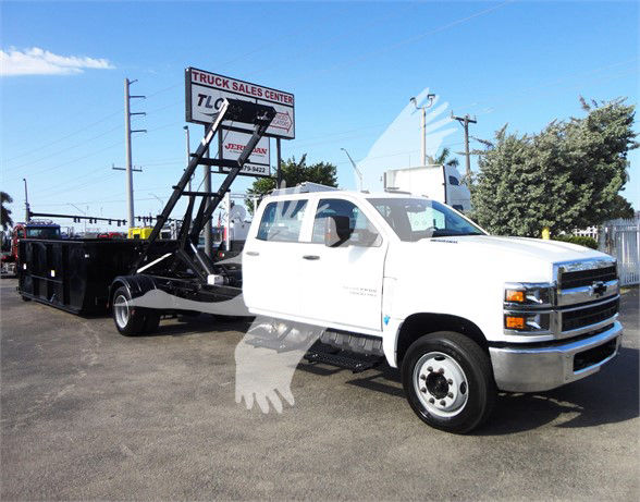 Delivery truck with a white Chevrolet Silverado 5500HD cab and a black gooseneck trailer in a dealership lot. Signage reads 'TRUCK SALES CENTER' and other text in background.