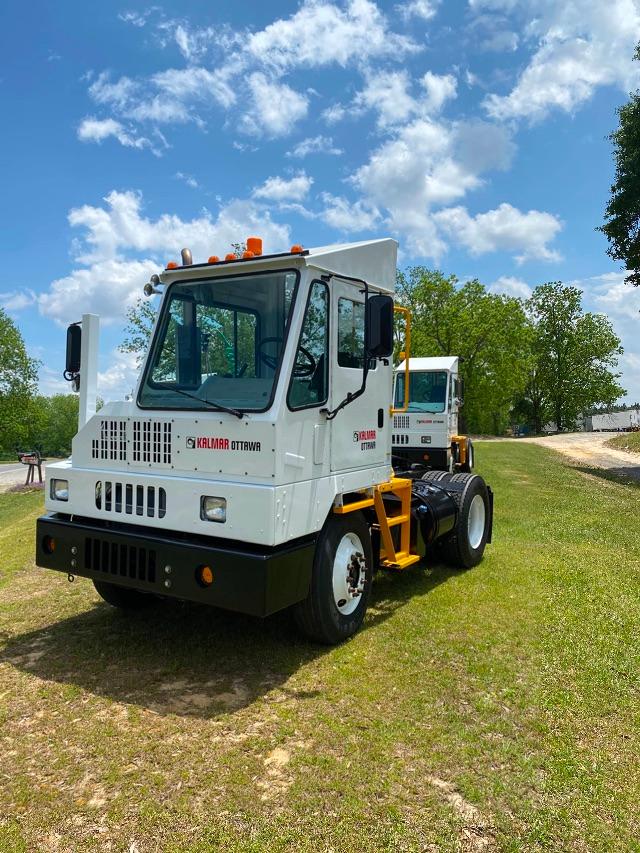 Tow tractor/yard truck showing white cab with Kalmar Ottawa branding, black bumper, orange accents, and large glass windows. Front view on grassy lot with another truck in background.