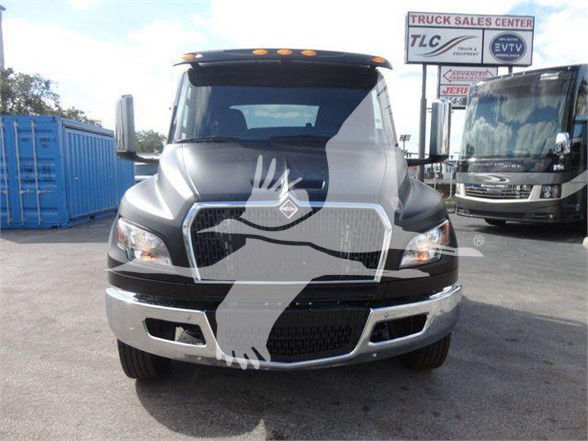 Front view of a 2024 International MV truck with a large chrome grille and bumper. Visible text on signs in background include Truck Sales Center TLC and EVT. No obvious model specifics on the truck body itself.