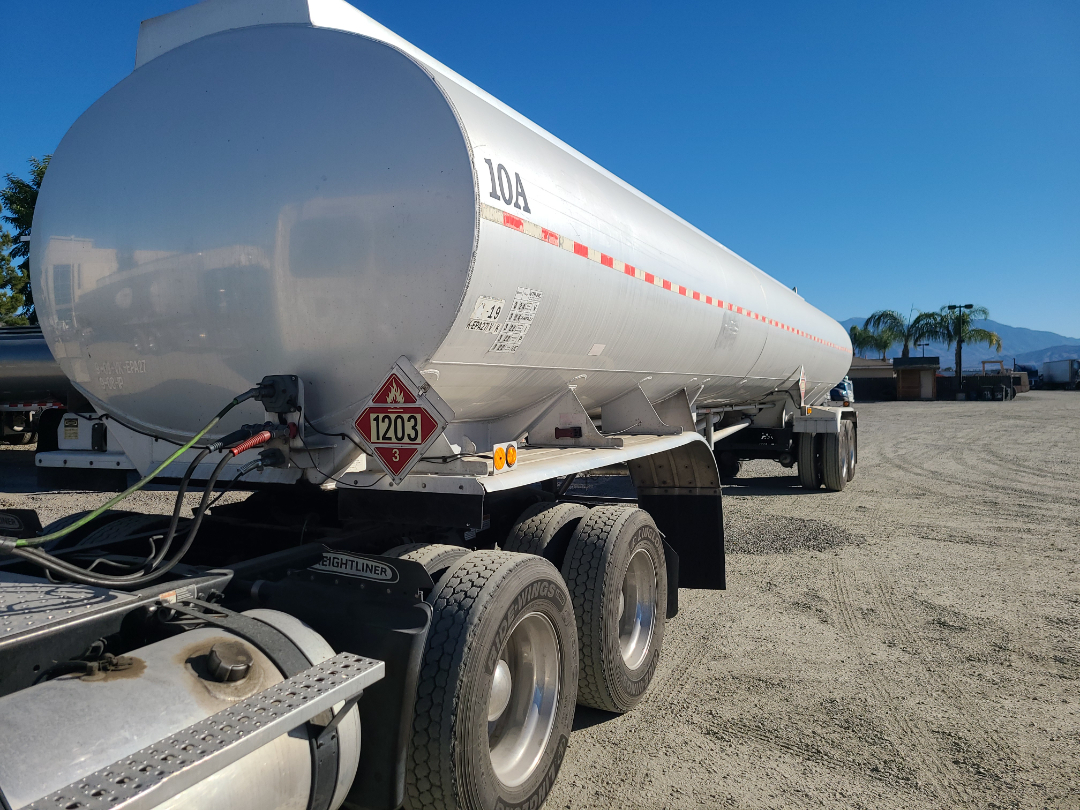 Trailer fuel tank semi-trailer with red dangerous goods placard 1203 and LOA marking, mounted on a multi-axle tractor in a gravel lot.