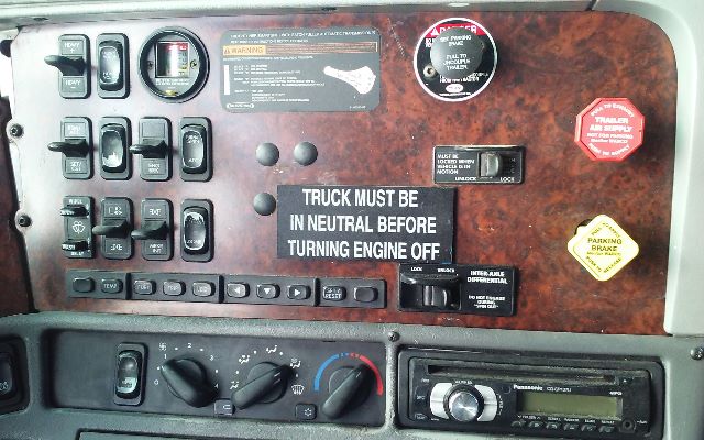 Dashboard and control panel of a Freightliner truck cab with various switches, warnings, and a central plaque reading 'TRUCK MUST BE IN NEUTRAL BEFORE TURNING ENGINE OFF.'