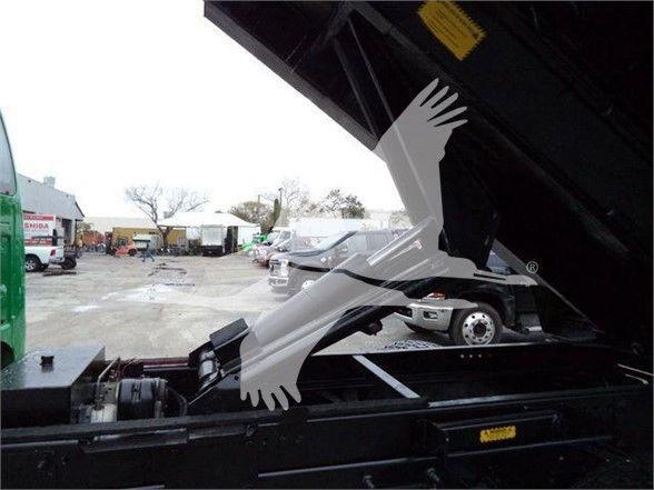 Dump truck bed area with hydraulic components; visible label on yellow warning tag and chassis; background shows other trucks and dealership lot.