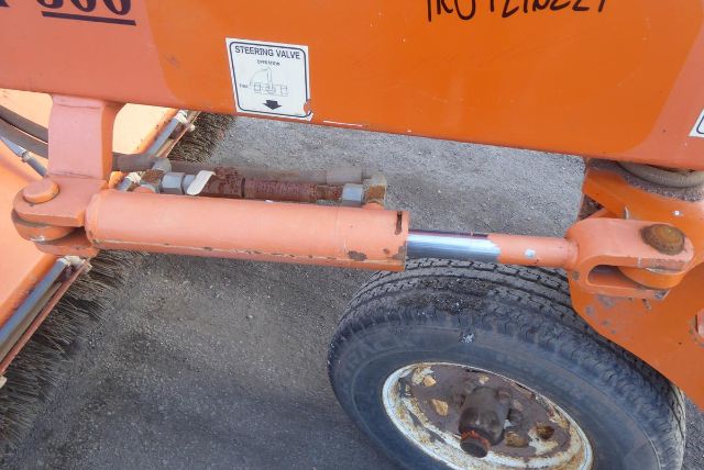 Trucking equipment trailer/axle showing hydraulic cylinder and wheel with tire; visible orange paint and steering valve sticker on the body.