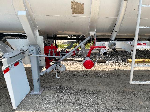 Trailer underside showing aluminum DOT-407 crude oil tank with red valve assemblies and piping, support legs, and mud/dirt ground.