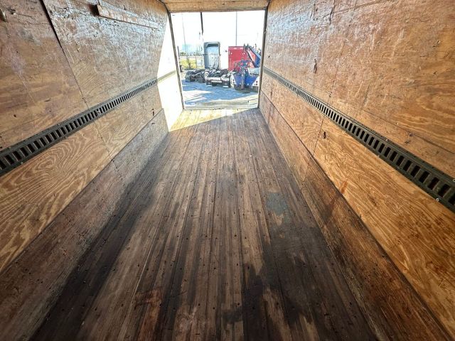 Interior of a box truck trailer with wooden walls and floor, long empty cargo area, rear doors open showing other trailers outside.