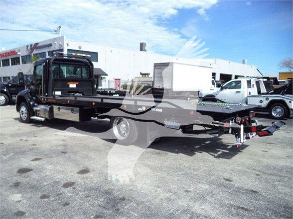 Kenworth T280 flatbed tow truck with extended trailer and equipment, side view showing front cab, long flatbed, and tow hitch in a dealership lot.