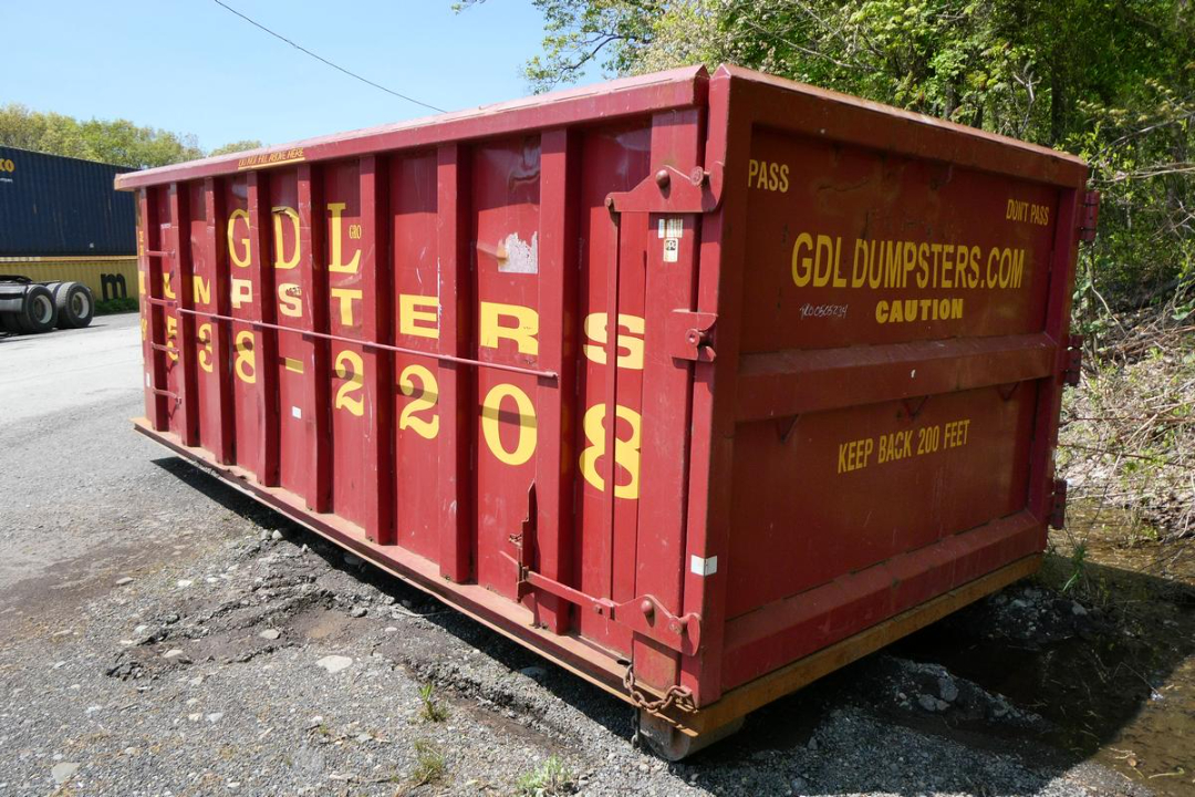 Red dumpster/container with yellow GDL DUMPSTERS lettering and Keep Back 200 Feet warning, side doors, on gravel surface.