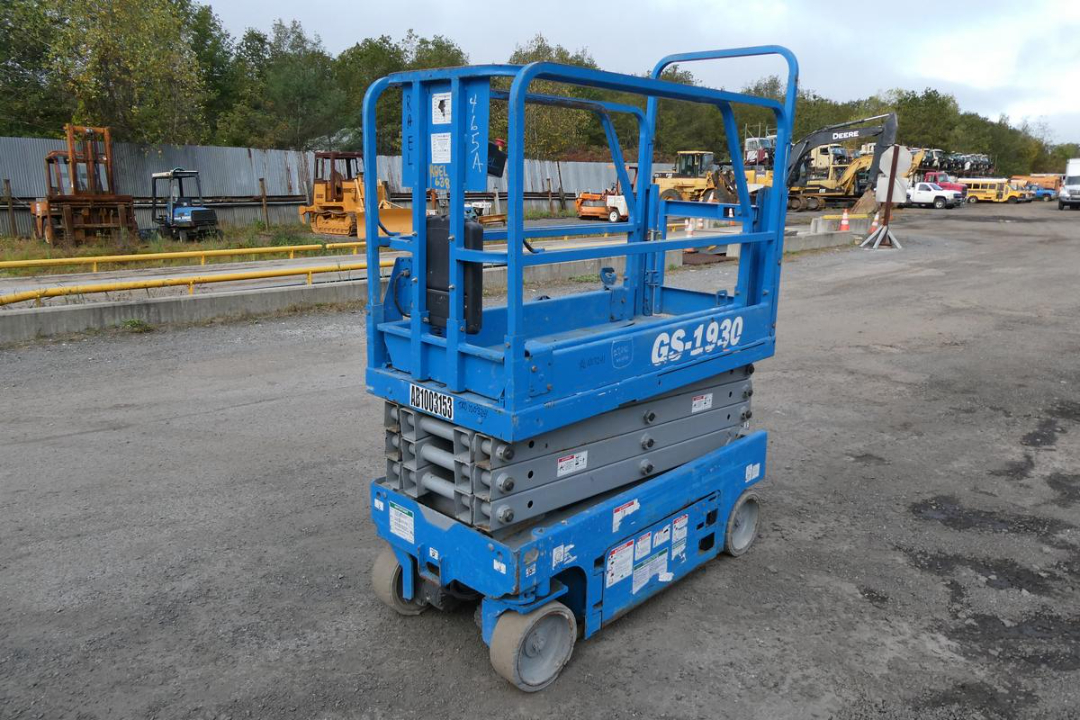 Blue Genie GS-1930 scissor lift with guard rails and gray hydraulic scissor mechanism, labeled GS-1930 on side, stacked on wheels in an outdoor lot.