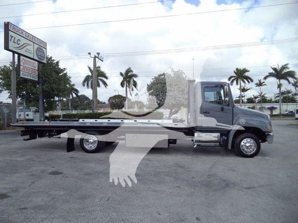 Flatbed tow truck profile view with Hino badge, long metal bed, and wheel lugs visible. Palm trees and dealership sign in background.