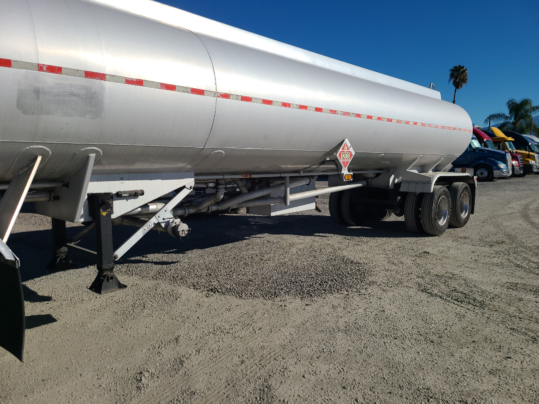 Rear side of a silver Beall fuel tanker trailer with tandem axles and a red-and-white hazard placard, parked on a gravel lot with other trucks in the background.