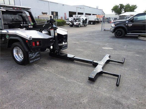 Tow truck with a metal hitch attachment on the ground, front of the truck visible with spare wheel, bumper guards, and black frame. Some text on the side of the truck reads TLC? and a phone number in white.